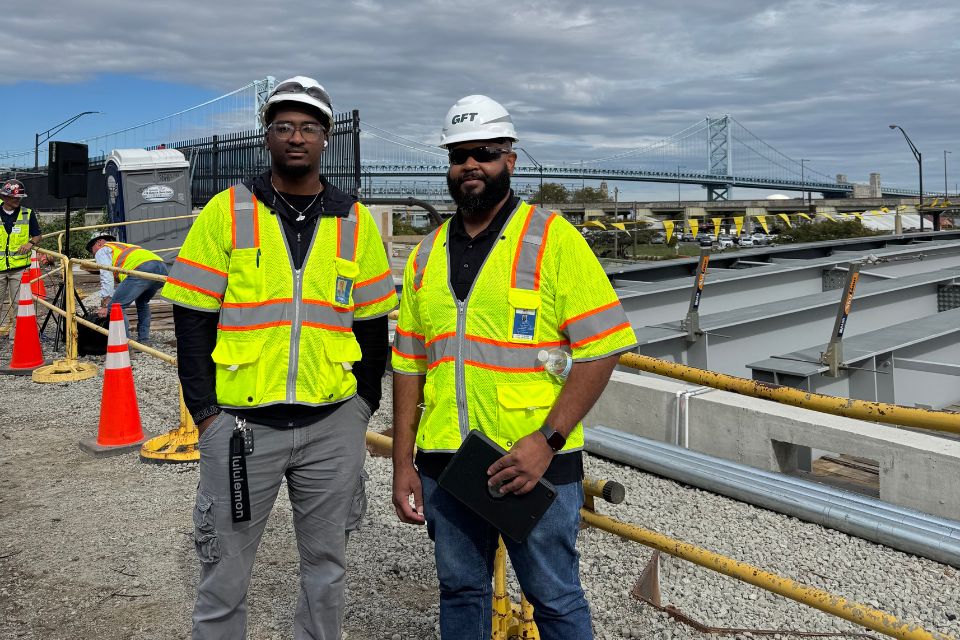 A student and alum stand in yellow vests and hard hats on a roof.