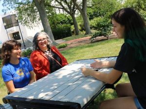 Two students and a faculty member sit at a picnic table with library in background