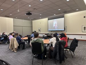 Students at tables watch as another student is speaking on screen
