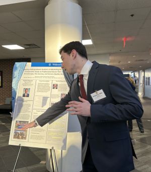 A student wearing a dark suit points and explains information on a poster