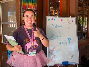 Sophia smiles at the camera giving a thumbs up. They wear a dark shirt and a pink tutu. Next to them is a white board with "Welcome to Brave Trails" written on it