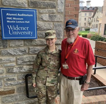 R-O-T-C cadet Meghan Rampolla, wearing fatigues, poses for a photo in front of Alumni Auditorium with her grandfather, Bob Hawley, wearing a P-M-C polo shirt and hat.