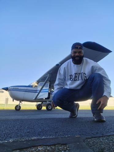 Delorean Andrews poses with an airplane. He wears a gray sweatshirt that reads "Boeing" and smiles at the camera.
