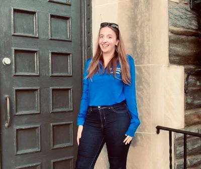 Zoe Sweet, wearing a blue shirt, stands in front of the door of Lathem Hall