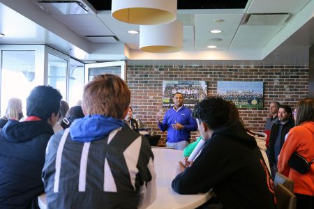 Students sit around a table listening to a professor talking