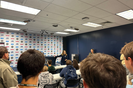 A woman wearing a dark blue shirt speaks to a crowd. The back of peoples heads is visible in the foreground.
