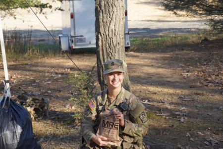 Meghan Rampolla in Army fatigues stands in a wooded area holding an M-R-E bag