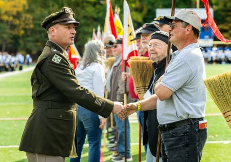 Lt. Col. Pascoe in dress uniform shakes the hand on a P-M-C alumnus holding a broom during the Homecoming Broom Drill.