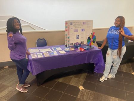 Two students stand at a table in the University Center covered in a purple tablecloth and info about Domestic Violence Awareness Month.