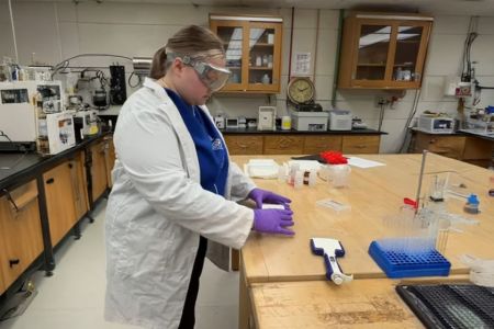 Bella Kuehn wears a lab coat and gloves while working in the chemical engineering lab.
