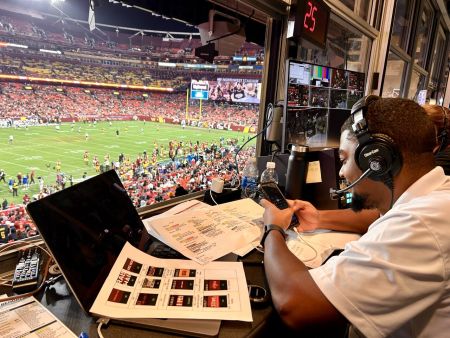 Andrew Proctor sits at a desk with a laptop and headphones on overlooking a football field