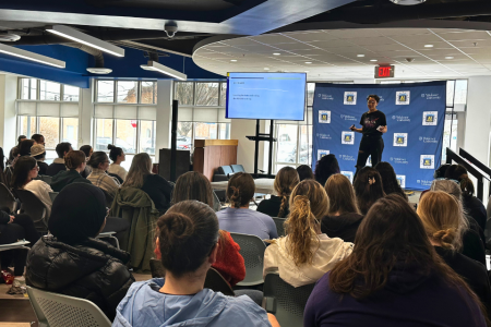 A student gives a presentation on a stage in front of a crowd. You see the back of the heads of members of the crowd, and the student is speaking and showing a presentation on a screen.