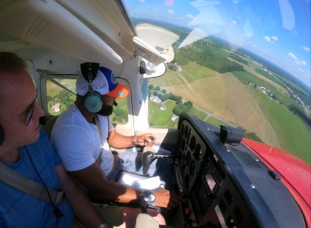 The view from the inside of an airplane cockpit. Delorean Andrews is piloting the plane and another man is in the seat beside him.