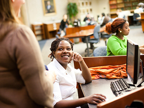 Student in library on computer and talking to another student